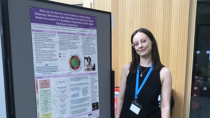 Lily standing with a research poster on sedentary behaviour when working from home.